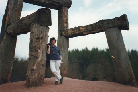 A woman wearing white and blue, standing at the foot of a giant wooden chair sculpture